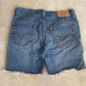 Levi's 501 jeansshorts - Klassiska blå jeansshorts från Levi's modell 501 med råa, fransiga benslut och patchad detalj på vänster ben 👖