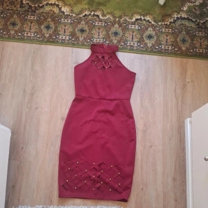Vinröd tight klänning med hög hals - Säljer en vinröd tight klänning med hög hals och dekorativa detaljer framtill. Klänningen har ärmlös design och snygga utskärningar med små guldfärgade nitar längst ner. Perfekt för festliga tillfällen.