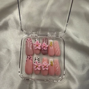 Rosa lösnaglar med blommor och djurmönster - Säljer ett set med rosa pres ons i olika designer. Naglarna har detaljer som 3D-blommor, gulddekorationer, zebramönster och leopardmönster. Flera nyanser av rosa och nude, vissa med glansig finish. Kommer i en genomskinlig ask. Och en nagelvårds sätt.