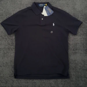Mörkblå pikétröja från Polo Ralph Lauren - Säljer en klassisk mörkblå pikétröja från Polo Ralph Lauren med vit broderad logga på bröstet. Tröjan har korta ärmar, krage och knäppning med två knappar. Perfekt för en stilren och sportig look.