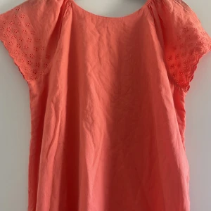 Korallfärgad bomullsklänning med broderade detaljer - Söt korallfärgad klänning i bomull med korta ärmar och broderade blommor på både ärmar och nederkant. Klänningen har en lös passform och är perfekt för varma dagar. Obs: taggen är kvar, aldrig använd 