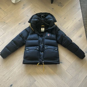 Svart dunjacka från Fjällräven Expedition Series - Säljer en svart dunjacka från Fjällräven Expedition Series med stor huva och gula detaljer. Jackan har dragkedja, knappar och två stora fickor framtill. Perfekt för kalla dagar och riktigt varm. Pris kan diskuteras 