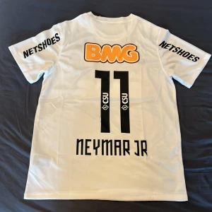 Santos tröja med Neymar Jr - En vit tröja med svarta detaljer på ryggen som har nummer 11 och namet Neymar Jr. Orangea och röda detaljer, klubbmärket och Nike swoosh loggan på framsidan. Luftig design perfekt för sommaren!