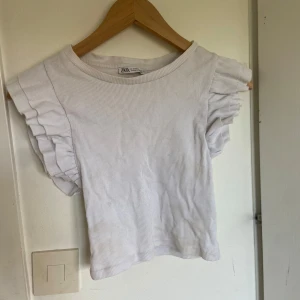 Vit ribbad topp med volangärmar från Zara - Säljer en söt vit ribbad topp från Zara med korta volangärmar. Toppen är i mjuk bomullsmix och har en croppad passform. Perfekt till sommaren och enkel att matcha med jeans eller kjol.