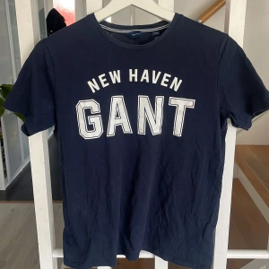 Mörkblå t-shirt från GANT med tryck - Säljer en mörkblå t-shirt från GANT med stort vitt tryck 'NEW HAVEN GANT' på bröstet. Klassisk rund halsringning och korta ärmar. Perfekt för dig som gillar stilrena och sportiga plagg.