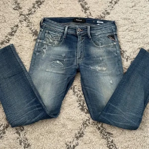 Blå Replay jeans med slitningar - Säljer ett par blå jeans från Replay med snygga slitningar och lätt tvättad look. Klassisk femficksmodell med silverfärgade detaljer och läderpatch bak i midjan. Perfekta för dig som gillar en avslappnad men trendig stil.