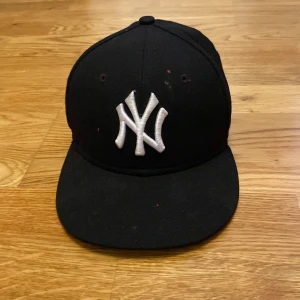 Svart New York Yankees keps - Klassisk svart keps från New York Yankees med vit broderad logga framtill. Modellen är en 59FIFTY från New Era och har en rak skärm. Storlek 7 (55,8 cm). Perfekt för dig som gillar sportig stil. Den har även fått lite färg i fram och därav blir den billigare 