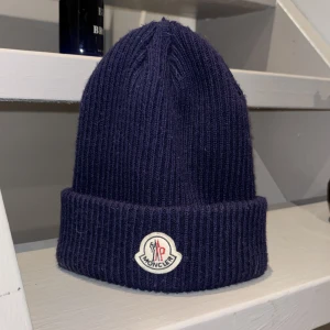 Moncler mössa  - Snygg mörkblå ribbstickad mössa från Moncler med uppvikt kant och klassisk logotyp framtill. Perfekt accessoar för kyliga dagar och enkel att matcha med olika outfits.