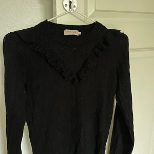 Svart stickad tröja med volang från Soaked in Luxury - Säljer en svart stickad tröja från Soaked in Luxury. Tröjan har lång ärm, rund halsringning och dekorativa volangdetaljer framtill. 