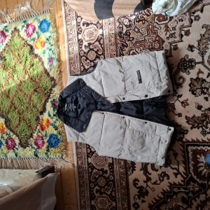 Beige dunväst från Canada Goose - Säljer en beige dunväst från Canada Goose med svart insida och hög krage. Västen har stora fickor framtill, dragkedja och knappar. Perfekt för lager-på-lager och håller dig varm under kyliga dagar.