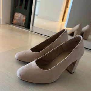 Beige/rosa mocka pumps med klack - Säljer ett par stilrena beige pumps från XIT i mocka-look. Skorna har rund tå och bred klack som ger stabilitet och komfort. Perfekta för dig som vill ha en klassisk och enkel sko med lite höjd. Perfekt till bal eller finmiddag. Använda inomhus en gång.