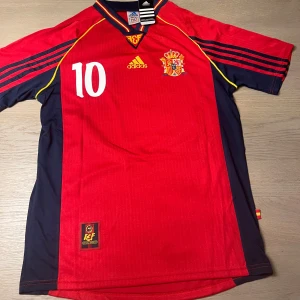 Spaniens landslagströja Raúl 10 Adidas - Säljer en klassisk röd Spanien fotbollströja med nummer 10 och Raúl på ryggen. Tröjan har gula detaljer, blå sidor och Adidas-logga samt landslagsemblem på bröstet. Kortärmad modell med sportig passform.