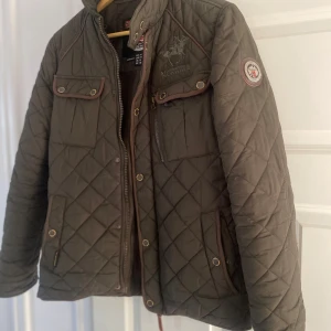 Olivgrön quiltad jacka från Geographical Norway - Säljer en olivgrön quiltad jacka från Geographical Norway med bruna detaljer och broderad logga på bröstet. Jackan har flera fickor med knappar och dragkedja samt patch på ärmen. Perfekt för kyligare dagar.