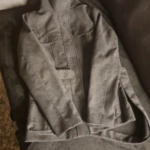 Grå kappa från H&M med bälte - Säljer en stilren grå kappa från H&M i storlek 40. Jackan har bälte i midjan, knappar på axeln och två bröstfickor. Fodret är ljusrosa och materialet känns mjukt och ulligt. Perfekt för kyligare dagar.