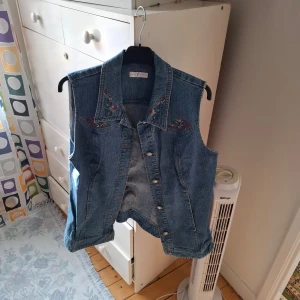 Blå jeansväst med broderi - Säljer en blå jeansväst med fina broderade blommor vid axlarna och kragen. Västen har knappar framtill och klassisk jeanskrage. Perfekt att styla över en t-shirt eller klänning för en cool look.
