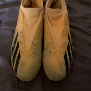 Gula fotbollsskor från Adidas - Säljer ett par gula fotbollsskor från Adidas med svarta ränder på sidorna och orange sula. Skorna har en slimmad design utan synlig snörning och är gjorda i syntetmaterial. Perfekta för dig som vill sticka ut på planen!