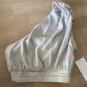 satin one-shoulder topp från NA-KD - Säljer denna snygga ljusbeige one-shoulder topp i satin från NA-KD. Aldrig använd och prislappen kvar på.