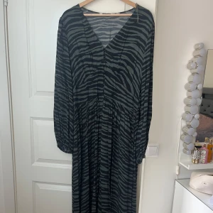 Zebramönstrad långklänning med v-ringning - Snygg långklänning med zebramönster i svart och grön. Klänningen har v-ringning och långa ärmar, samt ett avslappnat fall. Perfekt för dig som gillar djurmönster och vill sticka ut.