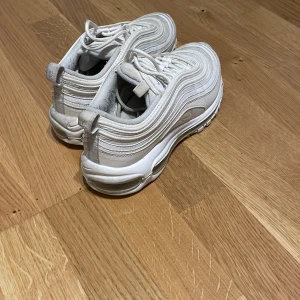 Nike Air Max 97 vita sneakers - Säljer ett par fräscha vita Nike Air Max 97 sneakers med den ikoniska vågiga designen och synlig luftbubbla i sulan. Skorna har snörning och är tillverkade i syntetmaterial med detaljer i mesh och läderlook.