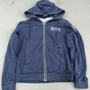 Blå hoodie från Hugo Boss - Snygg blå hoodie från Hugo Boss med vit broderad logga på bröstet. Hoodien har dragkedja framtill, huva med snörning och ribbade muddar. Perfekt för en avslappnad och stilren look.