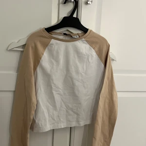 Beige och vit croppad långärmad tröja - Säljer en croppad långärmad tröja med vita paneler framtill och beige ärmar och krage. Perfekt för en avslappnad och trendig look. Tröjan har en enkel design utan synliga detaljer eller mönster.