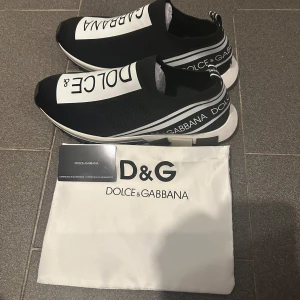 Svarta slip-on sneakers från Dolce & Gabbana - Säljer ett par svarta slip-on sneakers från Dolce & Gabbana med vit logga och detaljer. Skorna har en sportig design med elastisk ovandel och vit sula med svarta och grå inslag. Perfekta för dig som gillar stilrena och trendiga sneakers.