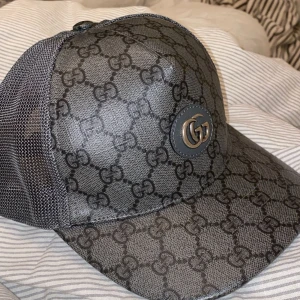 Grå keps från Gucci med GG-mönster - Snygg grå keps från Gucci med klassiskt GG-mönster över hela kepsen och ett GG-emblem framtill. Kepsen har mesh på sidorna och baksidan samt justerbart spänne i skinn baktill. Perfekt för dig som gillar exklusiva accessoarer.