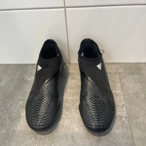 Svarta Adidas fotbollsskor med blå och röda detaljer - Säljer ett par svarta Adidas fotbollsskor med vita, blå och röda ränder på sidan. Skorna har en strumpliknande passform utan snörning och en mönstrad sula med blå detaljer för bra grepp på planen.