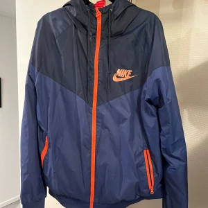 Blå och marinblå vindjacka från Nike - Snygg tvåfärgad vindjacka från Nike i blå och marinblå med orange detaljer och logga. Jackan har huva, dragkedja framtill och två fickor med dragkedja. Perfekt för en sportig look och enkel att matcha. Storlek L men sitter som M.