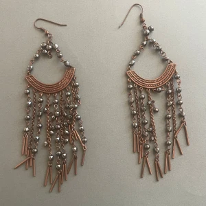 Stora bronsfärgade örhängen med kedjor och pärlor - Säljer ett par statement örhängen i bronsfärg med halvmåneformad detalj, kedjor och små metallpärlor. Örhängena har flera hängande kedjor med stavar och pärlor som ger en bohemisk och unik look.