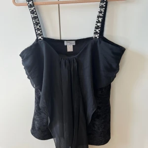 Svart sammetslinne med paljett- och pärlband från Heine - Säljer ett svart linnetopp från Heine i sammetsmaterial med breda axelband dekorerade med paljetter och pärlor. Toppen har en volangdetalj framtill i tunt tyg som ger en snygg effekt. Perfekt för dig som vill sticka ut lite extra.