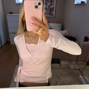 Ljusrosa omlottopp med knyt - Säljer en ljusrosa omlottopp med långa ärmar och knyt i midjan. Toppens material är mjukt och stretchigt, perfekt för en trendig och feminin look. Passar snyggt till både jeans och kjol. Supersöt färg och smickrande passform!