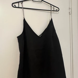 Svart klänning med kedjeaxelband - Säljer en svart klänning med smala kedjeaxelband och v-ringning. Klänningen har en enkel och stilren design som passar perfekt till festliga tillfällen. Materialet känns mjukt och följsamt mot kroppen.