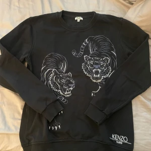 Svart sweatshirt med tigermotiv från Kenzo - Cool svart sweatshirt från Kenzo med två stora vita tigermotiv på framsidan och broderad Kenzo Paris-logga vid nederkanten. Tröjan har rund hals och långa ärmar, perfekt för en streetwear-look.