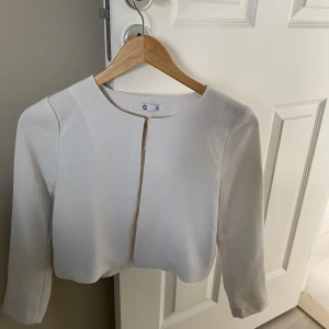 Vit croppad kavaj för barn - Stilren vit kavaj med croppad passform och rund halsringning. Jackan har lång ärm och stängs framtill med en diskret hake. Materialet är strukturerat och ger en elegant känsla. Perfekt för dig som vill ha en enkel men snygg look.