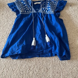 Blå blus med broderi från H&M - Säljer en blå blus från H&M med vita broderade detaljer upptill och knytband med tofsar. Blusen har korta ärmar och är tillverkad i ett lätt bomullsmaterial. Perfekt för varma dagar och ger en bohemisk känsla.