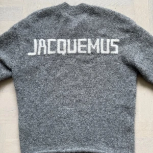 Grå stickad tröja från Jacquemus - Mysig grå stickad tröja från Jacquemus med vit logotyp på ryggen. Tröjan har rund halsringning och långa ärmar.                                               Nypris: 7800 kr                                                      Lite större i storleken