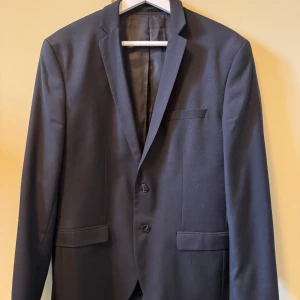 Mörkblå kostym från Jack & Jones - Säljer en stilren mörkblå kostym från Jack & Jones, modell Superslim Fit i storlek 50. Kavajen har två knappar och klassiska fickor, och byxorna har bakficka med knapp. Perfekt för bal eller fest. Kavajen är storlek 50 och byxorna är storlek 52. 