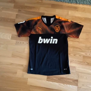Valencia CF fotbollströja Puma - Säljer en svart Valencia CF matchtröja från Puma med orange och svarta detaljer på axlarna. Tröjan har klubbmärke på bröstet och sponsorlogga i vitt framtill. Perfekt för dig som älskar spansk fotboll.