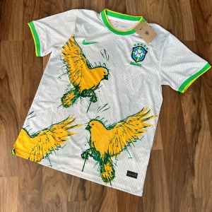 Brazil special edition - Säljer en vit Brasilien fotbollströja från Nike med gröna och gula detaljer. Tröjan har stora gula papegojor tryckta på framsidan, grönt och gult runt kragen och ärmslut samt Brasilien-emblem på bröstet. Tillverkad i Dri-FIT material.  Mått: Axel-Axel 44cm Armhåla-Armhåla 52cm längd 70cm
