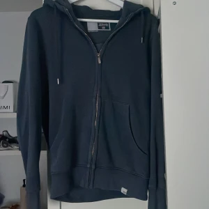 Mörkblå hoodie med dragkedja från Solid - Säljer en mörkblå hoodie från Solid i storlek XS. Tröjan har dragkedja framtill, huva med snören och en stor ficka på magen. Perfekt för en avslappnad stil och enkel att matcha med jeans eller joggers.