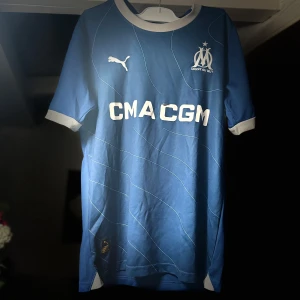 Mairseille tröja 23/24 - Snygg blå fotbollströja från Puma med vit logga och text. Tröjan har korta ärmar och en vit krage. Perfekt för fotbollsfans som vill visa sitt stöd. Den har en modern design med subtila linjer över hela framsidan.