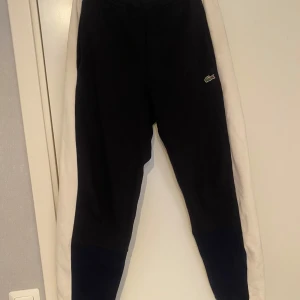 Mörkblåa mjukisbyxor från Lacoste - Mörkblå mjukisbyxor från Lacoste med vit sidopanel och klassisk broderad krokodillogga på benet. Regular fit och elastisk midja för skön passform. Perfekta för dig som gillar att chilla, är öppen för frågor är förhandling! Priset är inte hugget i sten