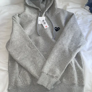 Grå hoodie från Comme des Garçons Play - Grå hoodie från Comme des Garçons Play med svart hjärtdetalj och ögon på bröstet samt stor hjärtdetalj på ryggen. Mjuk bomullskvalitet, huva med dragsko och känguruficka framtill. Perfekt för en avslappnad stil. Passar mer som en S men kan passa M! Priset kan prutas