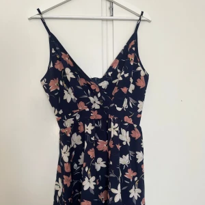 Mörkblå blommig klänning med smala axelband - Söt mörkblå klänning med smala axelband och blommigt mönster i vitt och rosa. Klänningen har v-ringad omlott-topp och utställd kjol, perfekt för varma dagar.