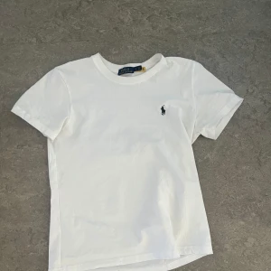 Vit t-shirt från Polo Ralph Lauren - Klassisk vit t-shirt från Polo Ralph Lauren med liten broderad logga på bröstet. T-shirten har rund halsringning och korta ärmar. Perfekt basplagg med stilren design och mjukt bomullsmaterial. En liten defekt som du ser på bild 2 men syns knappast. Storlek m men passar nog mer S