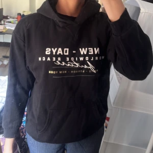 Svart hoodie med tryck  - Svart hoodie med vit och guld text på bröstet: 'NEW - DAYS WORLDWIDE REACH' och motiv på ryggen med leopard och text. Stor känguruficka framtill och huva. Perfekt för en chill och avslappnad stil.