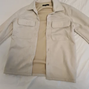 Beige overshirt från Smog - Säljer en stilren beige overshirt från Smog i storlek S. Jackan har en avslappnad passform med två bröstfickor och knappar framtill. Perfekt för lager-på-lager-stil under höst och vår.priset kan diskuteras vid snabbaffär 