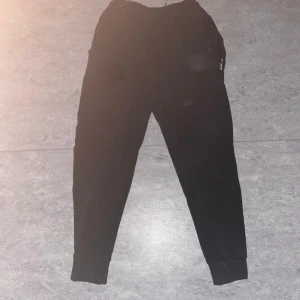 Nike Tech Fleece Byxor - Säljer ett par svarta Nike Tech byxor med snörning i midjan. Byxorna har en avslappnad passform och är perfekta för en bekväm stil. De har ribbade muddar vid bensluten och praktiska fickor på sidorna.