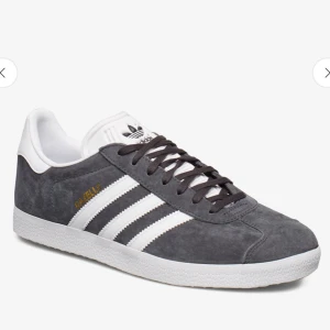 Grå Adidas Gazelle sneakers - Snygga grå Adidas Gazelle sneakers med vita detaljer och klassiska tre ränder på sidan. Skorna har snörning och är tillverkade i mocka för en stilren look. Perfekta för både vardag och avslappnade tillfällen. Dem har även på sig vita skosnören nu istället för svarta. Fint skick 